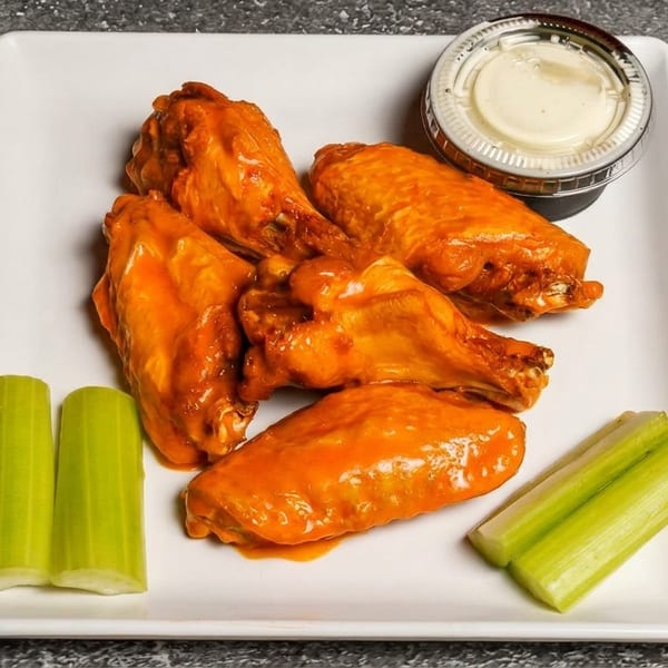 Five buffalo chicken wings with celery sticks and a side of ranch dressing on a white plate.