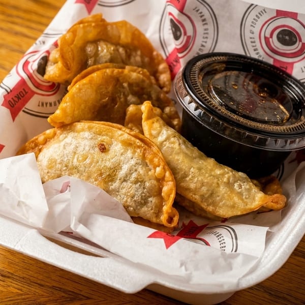 Fried dumplings and a container of dipping sauce in a takeout box lined with branded paper.